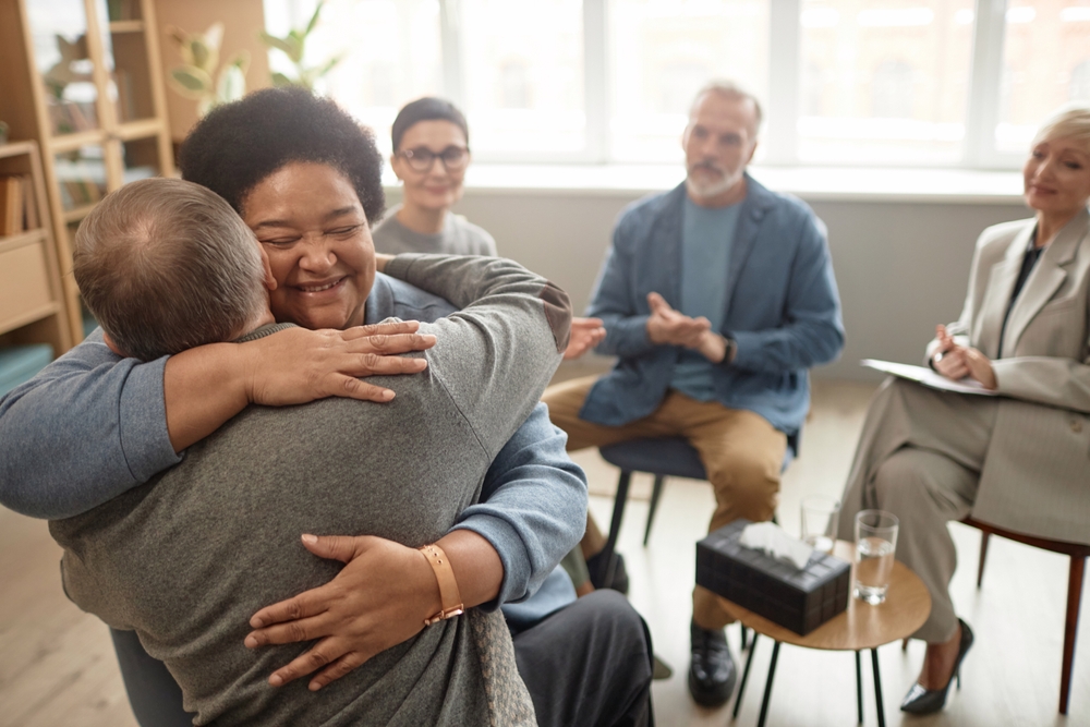 Portrait of Black senior woman embracing friend in group therapy session and smiling happily celebrating recovery copy space