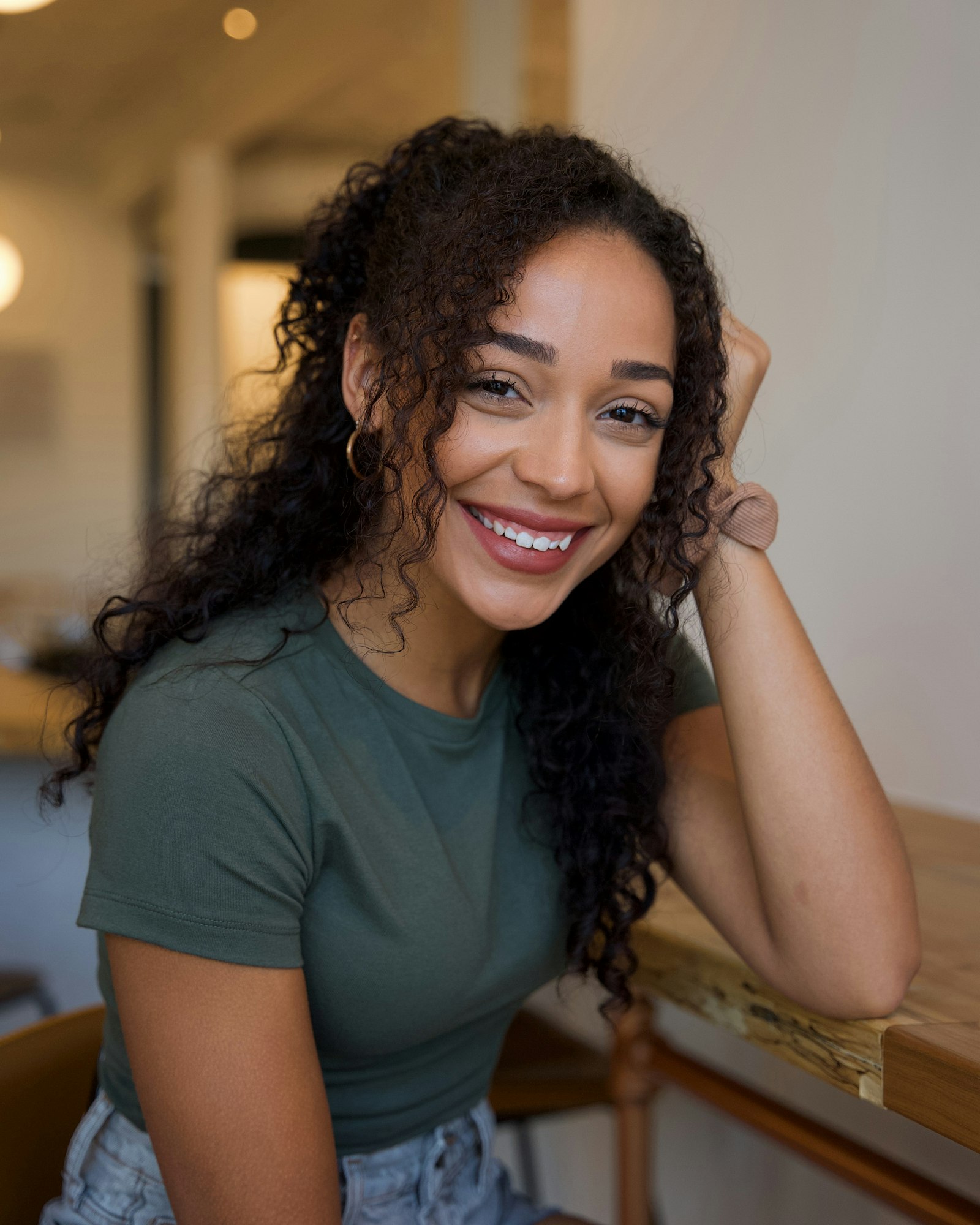 A young woman with curly hair smiling warmly while sitting at a wooden table